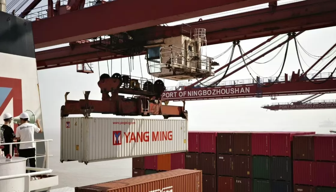 Ein Container wird im Hafen von Ningbo Zhoushan von einem Kran angehoben, während Arbeiter zuschauen.